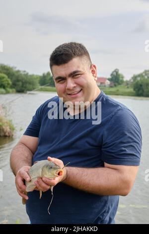 Der Fischer fing einen Karpfen. Kaukasischer junger Mann, der an einem sonnigen Sommertag Karpfen in den Händen hält. Ruhe auf dem Land, Camping Stockfoto