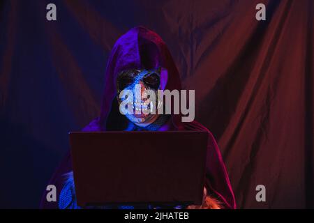Portrait eines Zombies in Hemd und Kapuzenumhang gekleidet mit seinem Laptop. Die Szene ist dunkel, beleuchtet von blauen und orangen Lichtern, das Gesicht des Monsters ist illumi Stockfoto