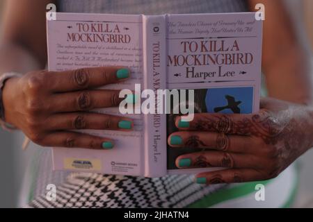 Schwarze Frauenhände mit Henna-Tattoo und Aqua-Nägeln halten das Buch auf, um Einen Mockingbird zu töten, auf Beinen in schwarz-weißen Hahnentritthosen. Stockfoto