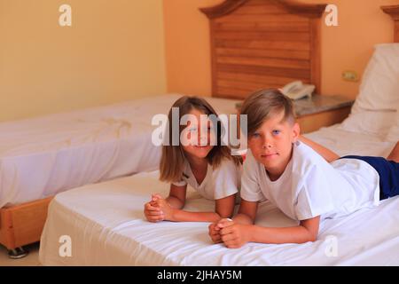 Zwei Kinder liegen im Urlaub in einem Hotelzimmer auf einem Bett mit weißen Laken. Kinder spielen zu Hause auf dem Bett mit weißen Laken. Junge und Mädchen, bevor sie zu Hause ins Bett gehen Stockfoto