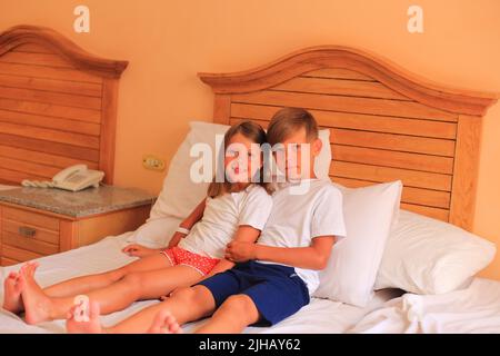 Zwei Kinder liegen im Urlaub in einem Hotelzimmer auf einem Bett mit weißen Laken. Kinder spielen zu Hause auf dem Bett mit weißen Laken. Junge und Mädchen, bevor sie zu Hause ins Bett gehen Stockfoto