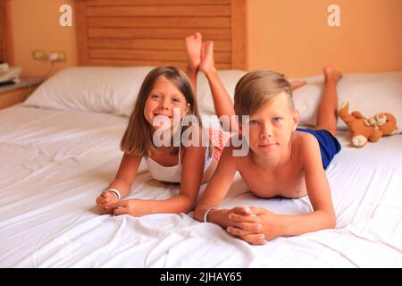 Zwei Kinder liegen im Urlaub in einem Hotelzimmer auf einem Bett mit weißen Laken. Kinder spielen zu Hause auf dem Bett mit weißen Laken. Junge und Mädchen, bevor sie zu Hause ins Bett gehen Stockfoto