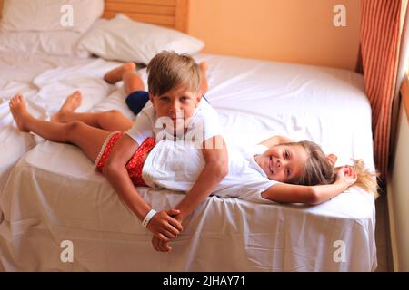 Zwei Kinder liegen im Urlaub in einem Hotelzimmer auf einem Bett mit weißen Laken. Kinder spielen zu Hause auf dem Bett mit weißen Laken. Junge und Mädchen, bevor sie zu Hause ins Bett gehen Stockfoto