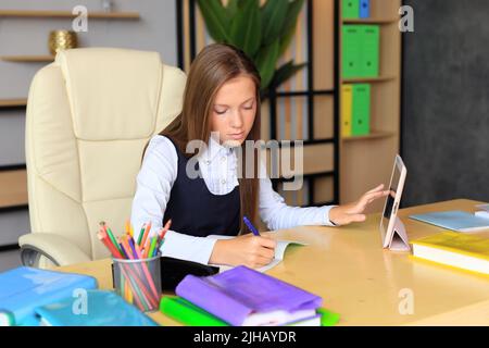 Ein Mädchen in Schuluniform sitzt am Tisch für den Unterricht. Das Kind macht Hausaufgaben mit Büchern und einem Tablet. Klassenzimmer Stockfoto