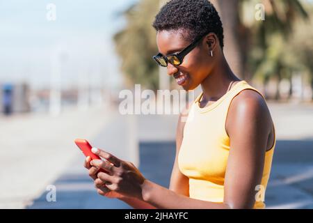 Trendige afroamerikanische Frau, die an einem sonnigen Sommertag ihr Telefon im Freien benutzt Stockfoto