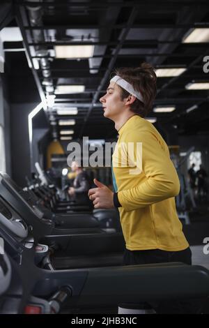 Vertikale Profilaufnahme eines männlichen Athleten mit Stirnband, der im Fitnessstudio auf dem Laufband läuft Stockfoto