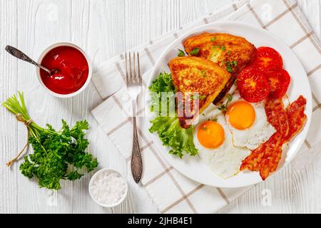 irisches Frühstück mit Kartoffelpüree, Spiegeleiern, Speckscheiben, gegrillten Tomaten und frischem Salat auf weißem Teller auf weißem Holztisch, horizontal Stockfoto