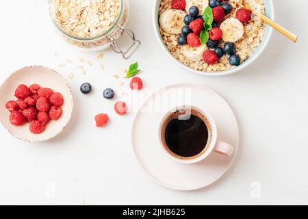 Kaffee und hausgemachter Haferbrei mit Himbeeren, Heidelbeeren, Banane und Chia-Samen auf weißem Tisch. Gesundes Frühstück. Draufsicht, flach liegend. Stockfoto