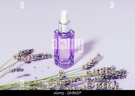 Kosmetisches Serum in Glasflasche und Lavendelblüten auf lilafarbischem Hintergrund. Aromatherapie und Naturkosmetik Konzept. Nahaufnahme. Stockfoto