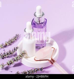 Hautpflegeprodukte, Serum- und Kosmetiköl, Rosenquarzkristall-Gesichtswalze und Lavendelblüte auf hellviolettem Hintergrund. Aromatherapie und natürlich Stockfoto