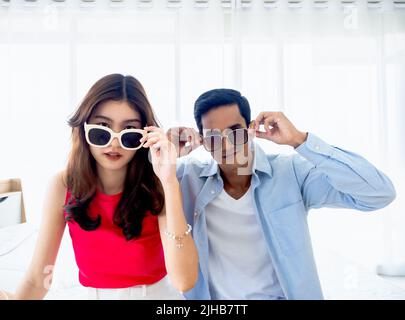 Glückliches asiatisches Paar im Sommer, junger Mann und Frau tragen eine Sonnenbrille, berühren sie und schauen auf Kamera auf dem Bett im hellen Schlafzimmer in der Nähe des Glases Stockfoto