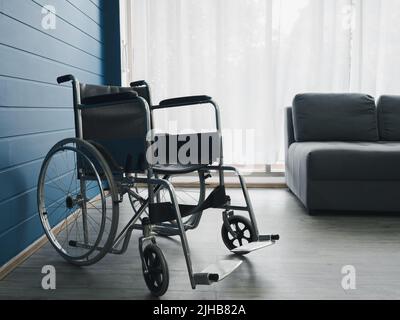 Leerer schwarzer Rollstuhl nahe der blauen Wand und grauer Sofasitz mit weißem Vorhang am Glasfenster im Wohnzimmer, der auf Patientendienste wartet. Stockfoto