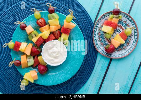 Frische Obstspieße auf einem blauen Teller serviert mit Joghurt Dip. Stockfoto