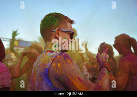 Der junge Kaukasier lächelt, als er Fotos und Videos von der Menge macht, die ebenfalls mit farbigem Pulver bedeckt ist, auf dem Color Walk in Dubai, Vereinigte Arabische Emirate. Stockfoto