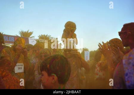 Das kleine Mädchen auf dem Huckepack bedeckt den Mund mit beiden Händen, während sie beim Color Walk in Dubai, VAE, die mit farbigem Puder umhüllte Menge untersucht. Stockfoto