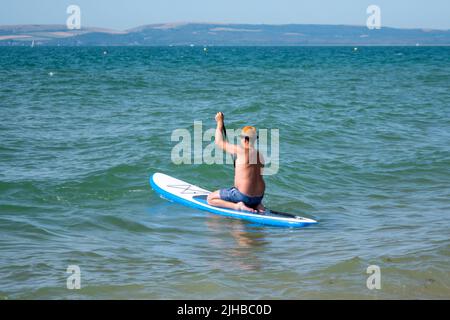 Boscombe Beach, Bournemouth, Dorset, England, Großbritannien, 17.. Juli 2022, Wetter. Über die Südküste zieht eine rekordverdächtige Hitzewelle ein, und vom Met Office wird eine bernsteinfarbene Warnung vor Hitze ausgegeben. Ein Paddlebarder paddelt in einem Schweinebutte-Hut ins Meer. Kredit: Paul Biggins/Alamy Live Nachrichten Stockfoto