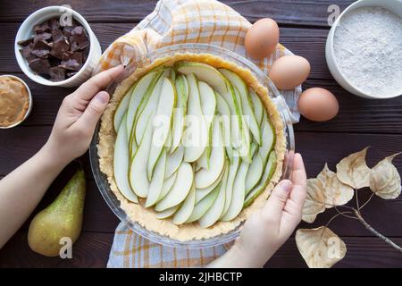 Die weibliche Hand hält den Birnenkuchen auf dem braunen Tisch, Draufsicht. Herbstlaub Dekor und Zutaten. Stockfoto