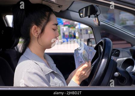 Nahaufnahme Porträt einer glücklich lächelnden attraktiven asiatischen Frau hält Geldscheine in ihrem Auto an der Tankstelle sitzen Stockfoto