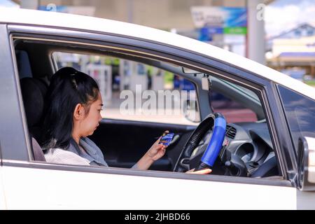 Junge, schöne asiatische Frau, die in ihrer Autohand sitzt und eine Kreditkartenzahlung an der Tankstelle hält Stockfoto