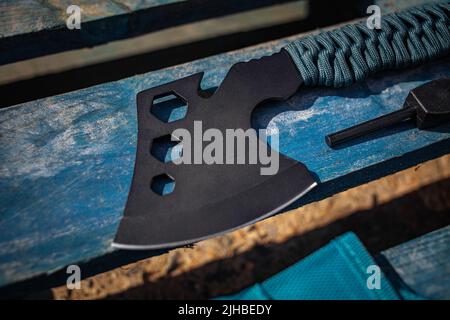 Eine kleine schwarze Axt mit einem Griff in Paracord-Verpackung. Ein Werkzeug zum Schneiden von Ästen oder Brennholz im Wald. Selbstverteidigungswaffe. Holzhintergrund. Stockfoto