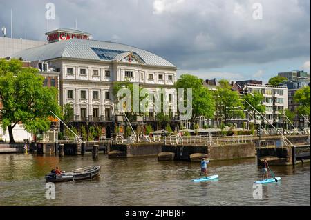 Boote, die an einem sonnigen Tag durch die Amstelsluizen Amsterdam fahren, mit dem Royal Theatre Carre im Hintergrund Stockfoto