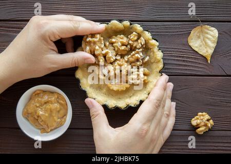 Leckere Tarte mit Nüssen und Karamellzubereitungen auf braunem rustikalem Hintergrund, Draufsicht. Weibliche Hände. Stockfoto