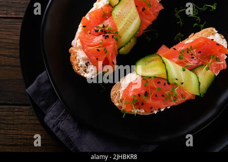 Sandwiches. Lachs-Toast mit Frischkäse, Gurke, schwarzem Sesam und Microgreens auf altem Holztisch-Hintergrund. Meeresfrüchte. Gesunde Ernährung. Fotografie Stockfoto