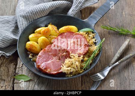 Deutsche Küche – geräucherter Schweinehalk mit Sauerkraut und gebratenen Babykartoffeln, die in einer eisernen Pfanne auf einem alten Holztisch serviert werden Stockfoto