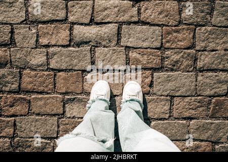Weibliche Beine in Jeans und weiße Sneakers auf Pflastersteinen. Die Füße im Teenageralter, Blick von oben. Prager Straße, Altstadt Stockfoto