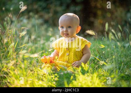 Kleine süße fröhliche Baby Mädchen auf dem grünen Gras an einem sonnigen Sommertag in gestrickten Baumwolle gelben Jumpsuit gekleidet sitzen. Stockfoto