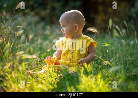 Kleine süße fröhliche Baby Mädchen auf dem grünen Gras an einem sonnigen Sommertag in gestrickten Baumwolle gelben Jumpsuit gekleidet sitzen. Stockfoto