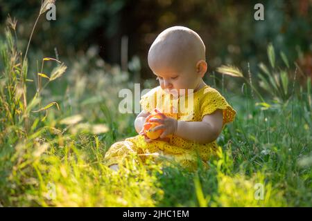 Kleine süße fröhliche Baby Mädchen auf dem grünen Gras an einem sonnigen Sommertag in gestrickten Baumwolle gelben Jumpsuit gekleidet sitzen. Stockfoto