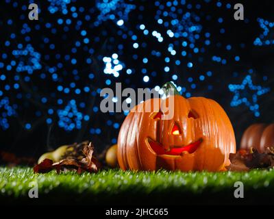 Großer orangefarbener halloween-Kürbis mit geschnitztem Gesicht, von innen beleuchtet, Früchte und Zapfen auf einem grünen Rasen unter einem blauen Sternenhimmel. Halloween An Feiertagen. Stockfoto