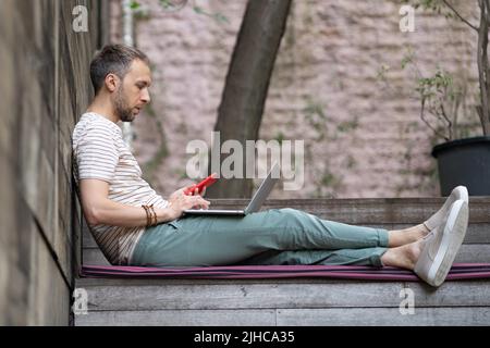Konzentrierter junger Unternehmer, der mit Laptop und Smartphone draußen sitzt, genießen Sie einen Arbeitstag unter freiem Himmel Stockfoto