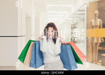 Porträt einer jungen lockigen schönen Frau, die in der Mall einkaufen geht und Kleidung kauft. Er hält farbige Papiertüten in den Händen, blickt lächelnd auf die Kamera Stockfoto