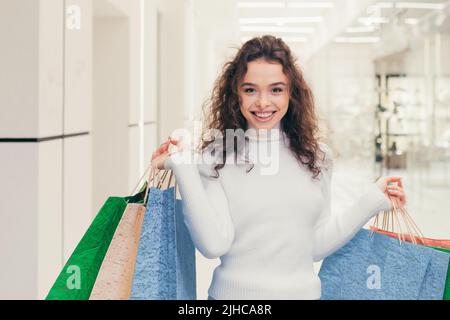 Porträt einer jungen lockigen schönen Frau, die in der Mall einkaufen geht und Kleidung kauft. Er hält farbige Papiertüten in den Händen, blickt lächelnd auf die Kamera Stockfoto
