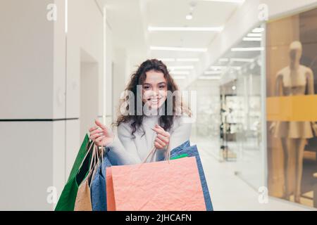Porträt einer jungen lockigen schönen Frau, die in der Mall einkaufen geht und Kleidung kauft. Er hält farbige Papiertüten in den Händen, schaut lächelnd auf die Kamera Stockfoto