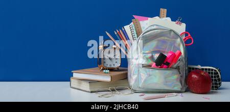 Schulrucksack mit Papeterie, Uhr und Apfel auf Tisch vor blauem Hintergrund Stockfoto