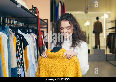 Eine junge, schöne Frau mit lockigen Haaren wählt, die in einem Bekleidungsgeschäft nach einer warmen gelben Jacke sucht, die sie auf einem Kleiderbügel an der Theke untersucht. Will kaufen, lächelt Stockfoto