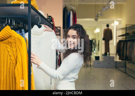 Junge schöne Frau mit lockigen Haaren wählt, auf der Suche nach einer weißen Jacke in einem Bekleidungsgeschäft, untersucht es auf einem Kleiderbügel auf dem Regal. Will kaufen, lächelt Stockfoto