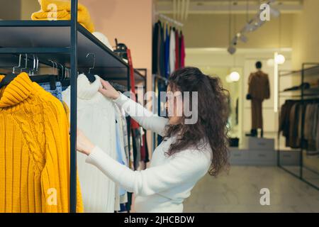 Junge schöne Frau mit lockigen Haaren wählt, auf der Suche nach einer weißen Jacke in einem Bekleidungsgeschäft, untersucht es auf einem Kleiderbügel auf dem Regal. Will kaufen, lächelt Stockfoto