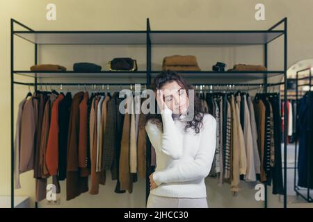 Beunruhigte und verwirrte junge Frau in einem Bekleidungsgeschäft in einem Einkaufszentrum, die ihren Kopf in der Hand hielt, überrascht von einer großen Auswahl. Steht auf dem Hintergrund der Regale mit Kleidung Stockfoto