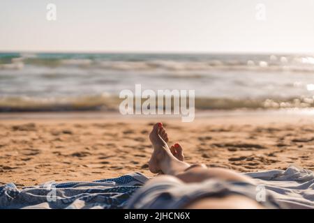 Die Füße einer Frau, die auf einem Handtuch am Strand in der Nähe des Meeres liegt Stockfoto