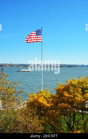 NEW YORK, NY - 04 NOV 2019: Fahnenmast mit Fallbäumen auf Ellis Island mit einem Statue Cruises Boot auf dem Fluss Stockfoto