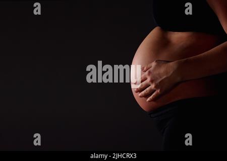 Schwanger Frau fühlt ihr Baby gegen schwarz - Copyspace. Mittelteil einer Schwangeren, die ihr Baby vor schwarzem Hintergrund fühlt - Copyspace. Stockfoto
