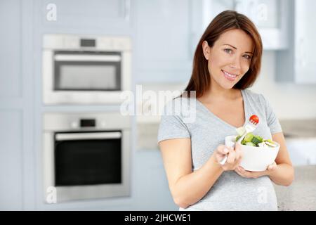 Eine gesunde Schwangerschaft zu garantieren. Porträt einer attraktiven erwarteten Frau, die einen gesunden Salat isst, während sie in ihrer Küche steht. Stockfoto