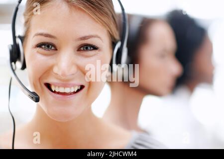 Wie kann ich helfen? Nahaufnahme einer hübschen Telefonanruferin, die im Hintergrund einen Anruf mit ihren Kollegen entgegennimmt. Stockfoto