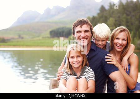 Genießen Sie den perfekten Familienurlaub. Lächelnde und glückliche junge Familie, die zusammen auf einem Steg sitzt. Stockfoto