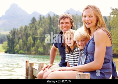 Genießen Sie die Zeit mit der Familie im Freien. Nette junge Familie, die am Steg in der Nähe eines Sees sitzt und lächelt. Stockfoto