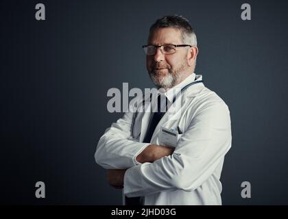 Ich bin der beste Chirurg auf meinem Gebiet. Studioporträt eines hübschen reifen männlichen Arztes, der mit gefalteten Armen vor einem dunklen Hintergrund steht. Stockfoto